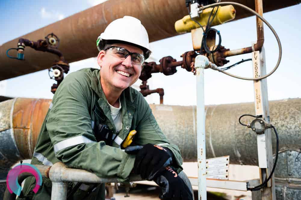 Smiling man in green overalls, hard hat, safety glasses and gloves standing underneath a large rusted pipe on a sunny day with blue sky