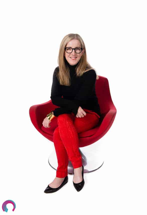 Corporate portrait of a female Brisbane executive wearing red pants and a black shirt, seated on a red chair, facing forward, leaning into the camera