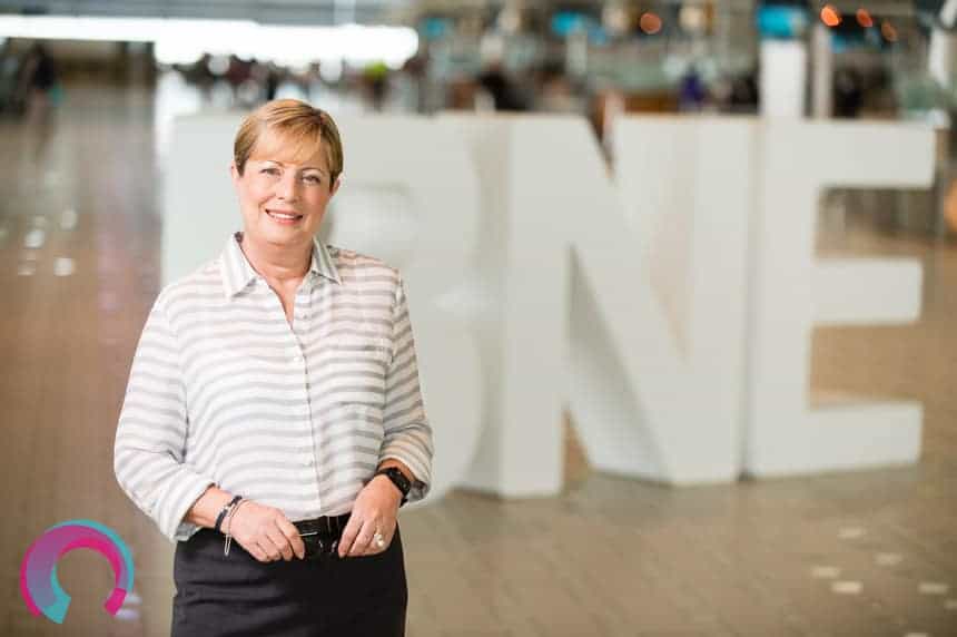 Corporate portrait of Julieanne Alroe, standing in the International Terminal Brisbane Airport, with three giant letters behind her. Letters are 'BNE"