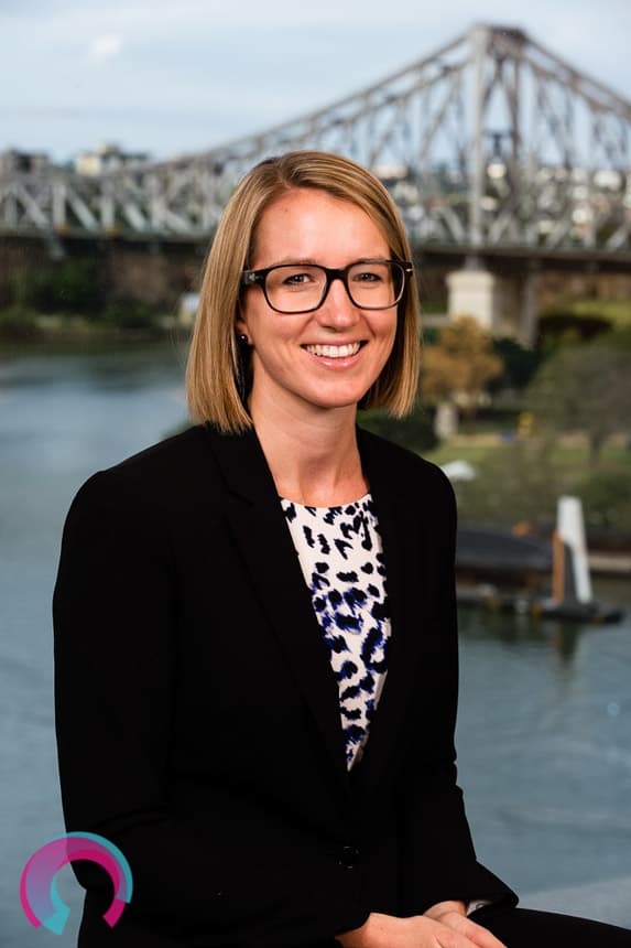 Corporate portrait of a blonde female with glasses sitting down, with Brisbane's Story Bridge as the background