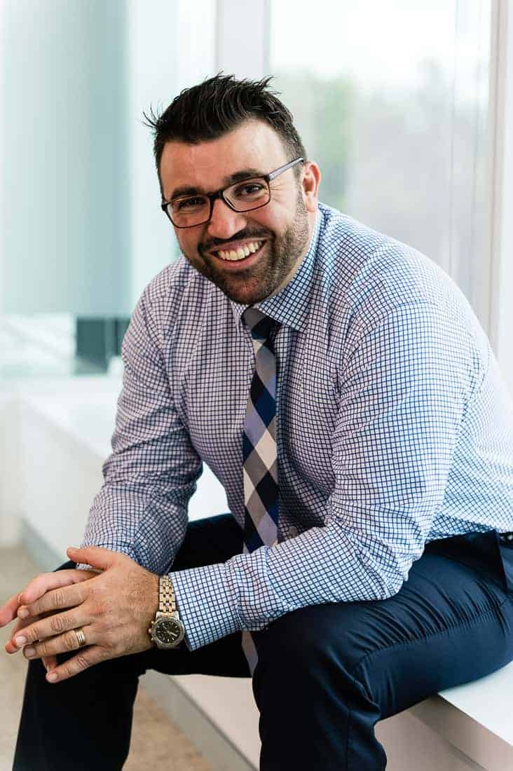 Portrait of a bearded man in a shirt and tie, sitting on a low window sill, leaning forward, smiling