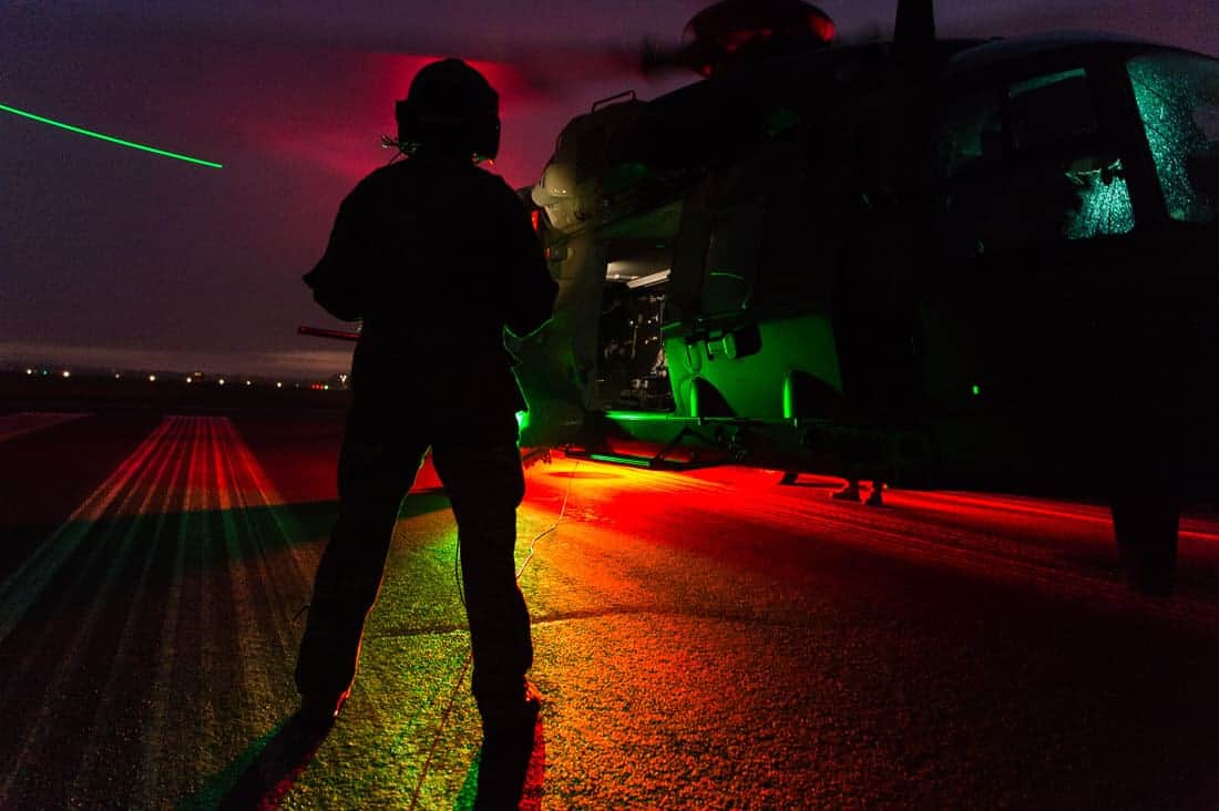 At dusk you see the silhouette of an MRH90 military helicopter from the side. In front of the helicopter you can see an aircrewman with flight suit and helmet standing with their back to the camera. The red and green ground lights of the MRH are visible, casting a green and red glows on everything. The piano keys (white landing strips of the runway) are visible beneath the MRH. The green tip lights of the helicopter are visible and we can see that the rotor blades are turning, as there is a trail of light caused by the movement of the rotor blades through the air